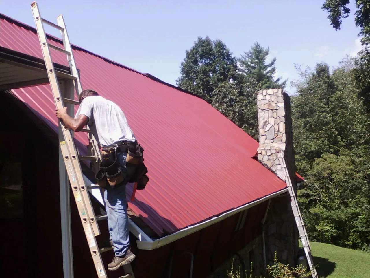 Roofer on a ladder beside a red metal roof and stone chimney — Soffit & Fascia Repair by Roofing Brothers in Logsden